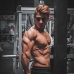 Muscular man posing in a gym, showcasing defined abs and biceps, representing fitness and bodybuilding in the context of steroid use and muscle enhancement.